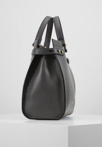 Musta nahkakäekott tekstuuriga külgpaneeliga, kahekordsete ülemiste käepidemetega ja kuldtooniste pandlatega, seisev valgel pinnal.