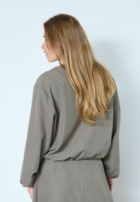 Hellgraues Kleid aus glattem Stoff mit lockerem Schnitt, langen Ärmeln und einem Taillenband, von hinten gesehen. Lange, wellige Haare sind sichtbar.