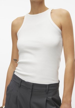 Hvid ribbet tanktop med høj hals, med en tætsiddende pasform. Bæres med skræddersyede grå bukser; synlig tatovering på armen.