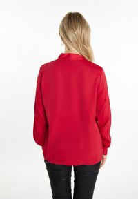 Rød satinbluse med krave og lange ærmer, der har en glat tekstur og en lige kant, parret med sorte tætsiddende bukser.