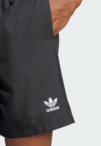 Schwarze Sportshorts aus glattem Stoff mit einem gestickten weißen Adidas-Logo nahe dem Saum. Sichtbare Seitenasche.