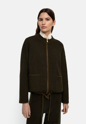 Kobieta ubrana w ciemnooliwkową kurtkę na zamek z przednimi kieszeniami na białą bluzkę z wysokim kołnierzem, w komplecie z dopasowanymi spodniami, na jednolitym tle.