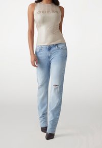 Top smanicato beige a costine con logo "GUESS", abbinato a jeans a gamba dritta strappati blu chiaro e décolleté nere a punta.