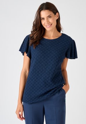Mujer con cabello castaño que lleva una camiseta texturizada azul marino de manga corta y pantalones azules marinos, sonriendo con una mano en el bolsillo.