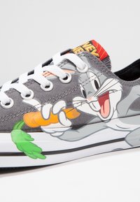 Sneaker in tela grigia con una grande grafica di Bugs Bunny che tiene una carota, lacci bianchi e una linguetta a motivo rosso e giallo.