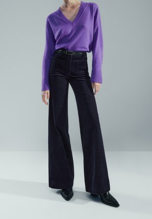 Person trägt einen violetten Pullover mit V-Ausschnitt, der in eine hoch taillierte, weite schwarze Hose mit Gürtel und schwarze spitze Schuhe gesteckt ist.