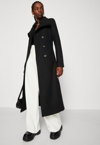 Lungo cappotto nero con colletto alto, bottoni doppi e silhouette pulita, abbinato a pantaloni bianchi a gamba larga e scarpe piattaforme nere.