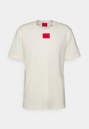 Crèmekleurig katoenen T-shirt met korte mouwen, voorzien van een rode rechthoekige label met de tekst "HUGO" in het midden. Zachte textuur, casual ontwerp.