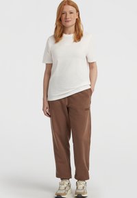 Camiseta blanca de manga corta, pantalones de chándal marrones de corte relajado con bordado del logo y zapatillas beige con acentos texturizados.