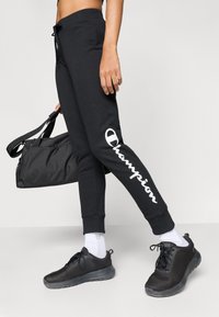 Μαύρα βαμβακερά παντελόνια jogging με λευκό λογότυπο «Champion» στο αριστερό πόδι, σετ με μαύρα παπούτσια από δίχτυ και μαύρη τσάντα duffel.