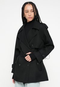 Czarny trencz z dwurzędowymi guzikami, szerokim kołnierzem i paskiem w talii. Charakteryzuje się gładką fakturą i płynnym krojem.