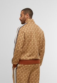 Brown patterned track jacket with high collar, featuring blue stripes on sleeves and a brown ribbed hem. Visible logo design throughout.