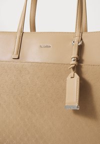 Beige handbag with a textured patterned lower section, smooth upper section, and logo detail; features dual handles and a detachable tag.