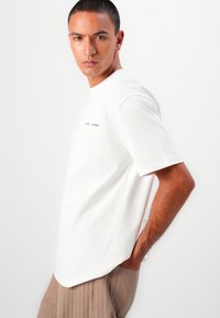 T-shirt en coton blanc à manches courtes, arborant un petit logo bleu imprimé sur la poitrine et une coupe décontractée, associé à un pantalon beige plissé.