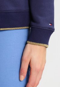 Sweatshirt marine avec des poignets côtelés ornés d'une bordure dorée métallisée ; petit détail de logo sur le poignet ; associé à un tissu bleu clair.