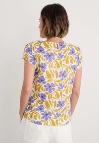 Top cu imprimeu floral în galben și violet pe un fundal alb, având mâneci scurte, un decolteu în formă de cheie la spate și o croială lejeră.
