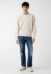 Sweat-shirt beige clair avec col rond, manches longues et poignets côtelés, associé à un jean bleu droit et des baskets blanches.