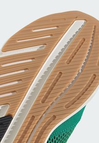 Podrážka topánky s textúrovaným hnedým gumovým vzorom, biela medzipodrážka a zelený vrchný materiál, ktorý má ventilačné otvory a kontrastné čierne detaily.