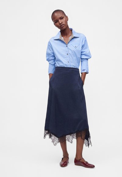 Femme portant une chemise rayée bleue boutonnée, une jupe midi bleu marine avec un ourlet en dentelle, et des chaussures Mary Jane bordeaux, debout les mains dans les poches.