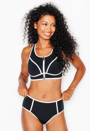 Donna con capelli ricci sorridente, indossa un bikini bianco e nero con cerniera frontale, aggiusta la parte inferiore con entrambe le mani.