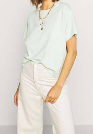 Femme portant un t-shirt Levi's vert pâle, un pantalon blanc taille haute et des colliers dorés superposés sur un fond uni.