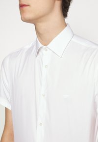 Muž v bílé košili na knoflíky s krátkým rukávem a špičatým límcem, viditelným horním trupem a krkem, otočený vlevo.