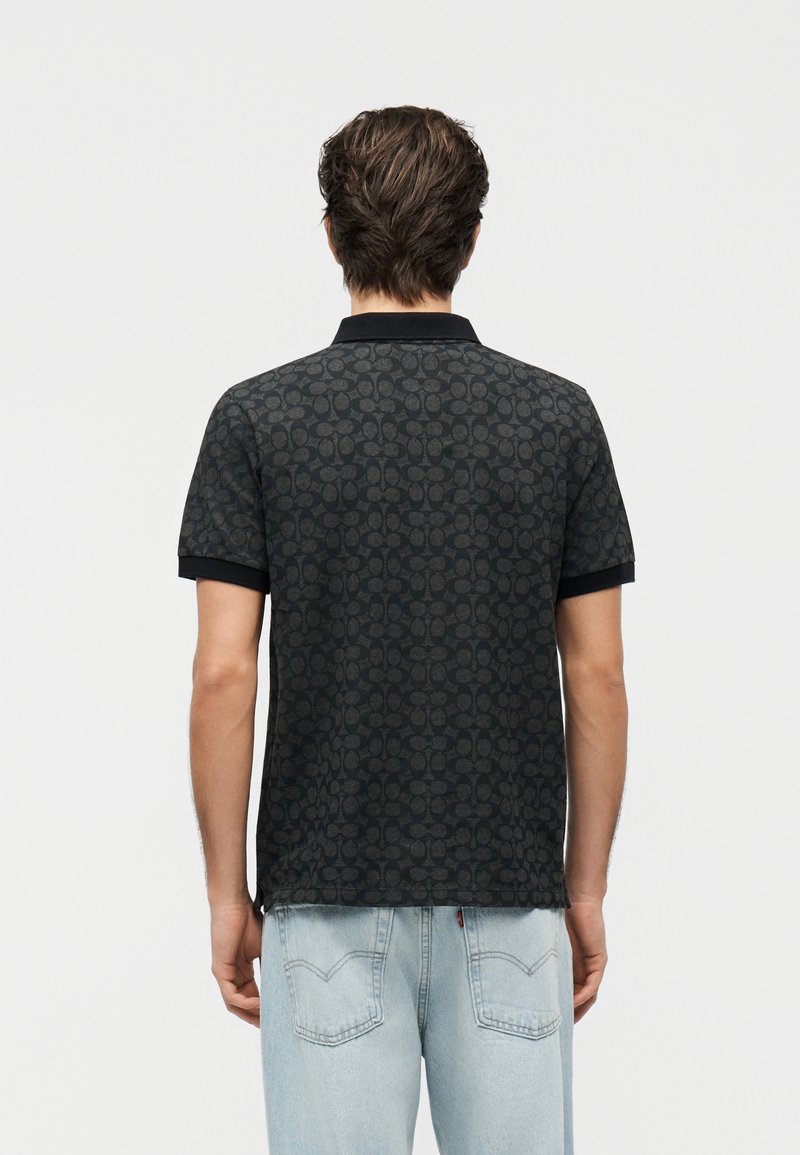 Homme portant un polo à manches courtes noir à motifs et un jean bleu clair, vu de dos sur un fond uni clair.