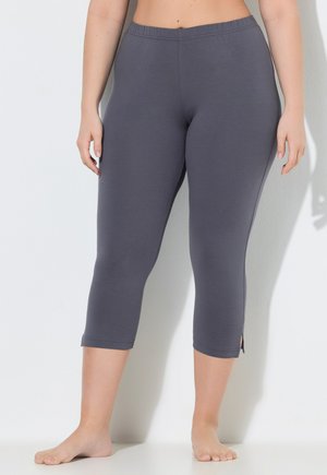 Kobieta w ciemnoszarych legginsach z wysokim stanem i małymi rozcięciami po bokach, stoi boso przy białej ścianie.