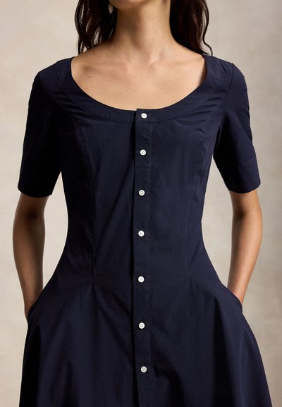 Γυναίκα που φοράει φόρεμα σε ναυτικό μπλε χρώμα με κοντά μανίκια, στρογγυλή λαιμόκοψη, λευκά κουμπιά μπροστά και τα χέρια στις πλαϊνές τσέπες.