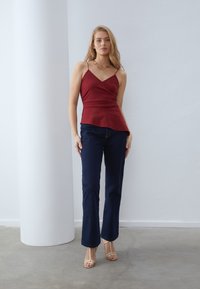 Burgundijska majica na bretele s omotanim dizajnom, u kombinaciji s tamnoplave wide-leg traperice i bež sandale s trakama. Glatka tekstura i minimalni detalji.