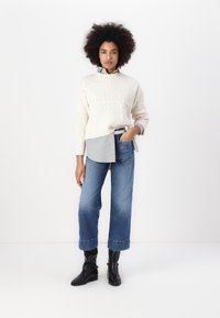 Pull en tricot blanc sur une chemise rayée bleu clair, associé à un jean bleu à jambes larges et des bottines noires. Look décontracté et superposé, propre.