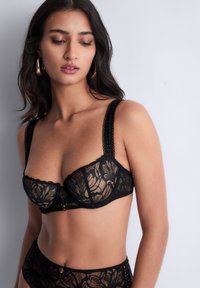 Soutien-gorge en dentelle noire avec des motifs floraux, support à armatures, larges bretelles texturées, et un détail en or au centre.