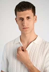 Λευκό πλεκτό πόλο με σχέδιο καλωδίου, κοντά μανίκια και γιακά. Το μοντέλο ρυθμίζει τον γιακά με ουδέτερο φόντο.