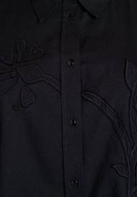Zwarte shirt met een knoopsluiting, geborduurd bloemenpatroon en subtiele kralenaccenten op de borduursels. Soepele stoftextuur.