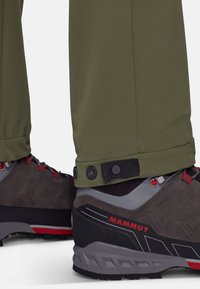 Olivengroene outdoorbroek met verstelbare boorden, gecombineerd met grijze wandelschoenen met rode accentkleuren en een gestructureerd oppervlak.
