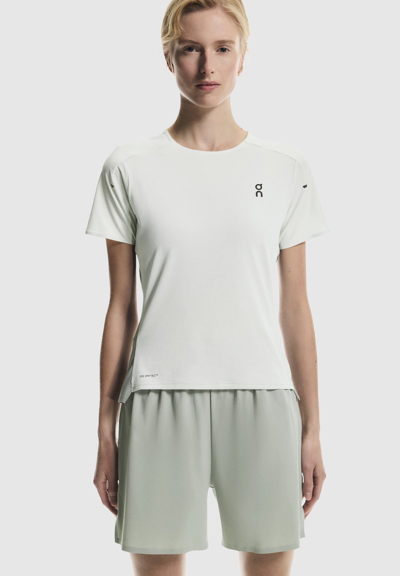 Jeune femme portant un T-shirt de sport blanc à manches courtes et un short gris clair, debout devant un fond uni gris clair.