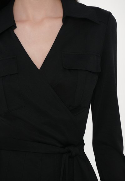 Čierne oblečenie vo forme šiat s golierom, dvoma prednými vreckami a opaskom v páse. Vyrobené z hladkej tkaniny s čistým, štruktúrovaným dizajnom.