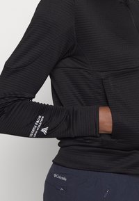 Maglietta nera a maniche lunghe con una texture a coste, caratterizzata da una zip al collo e un dettaglio del logo sul braccio sinistro. Il tessuto sembra leggero ed elastico.