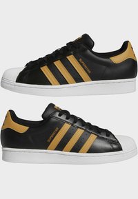 Black leather sneakers with three yellow stripes, white rubber toe cap, and rubber sole. "SUPERSTAR" logo featured on the side and heel.