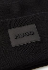 Svart strikket lue med teksturert overflate og et rektangulært, mørkt merke med hevet tekst "HUGO." Standard oppbrett-design.