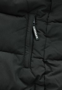 Čierna podložená bunda s vreckom na zips. Tkanina má hladkú textúru s jemným leskom. Štítok "Superdry" je viditeľný pri zipe.