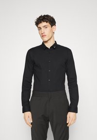 Camisa negra de manga larga abotonada con cuello estructurado, que presenta un sutil detalle cerca del cuello y un acabado de tela suave.