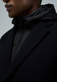 Abrigo de lana negro con un cuello estructurado, superpuesto a una chaqueta acolchada negra con cremallera, que presenta una textura suave y un diseño ajustado.