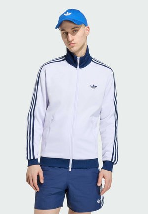 Junger Mann trägt eine weiße und marineblaue Trainingsjacke, marineblaue Shorts und eine blaue Adidas-Kappe, steht vor einem schlichten, hellen Hintergrund.