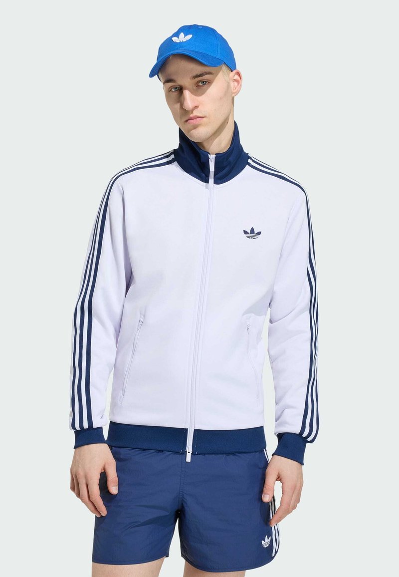 Joven con chaqueta deportiva blanca y azul marino, pantalones cortos azul marino y gorra azul de Adidas, de pie frente a un fondo claro y liso.