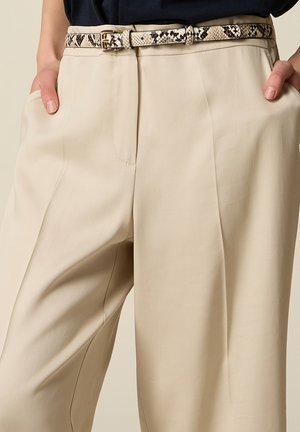 Pantalones beige a medida con pinzas delanteras y bolsillos laterales, usados con un cinturón fino con estampado de piel de serpiente y manos descansando en los bolsillos.