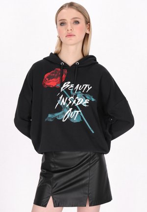 Junge Frau trägt einen schwarzen Kapuzenpullover mit einer roten Rose und dem Schriftzug „Beauty Inside Out“ kombiniert mit einem schwarzen Lederrock.