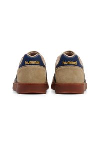 Beige suede sneakers with navy accents, featuring a round toe, thick brown rubber sole, and "hummel" logo in gold on the heel.