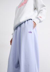 Svetložlté tepláky s elastickým pásom, zdobené vyšitou „nike“ nášivkou v levanduľovej farbe. Mäkká textúra, široký strih a bočné vrecká.