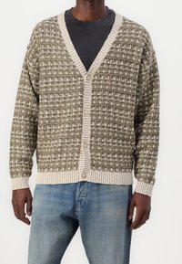 Stickad kofta i olivgrönt med vitt mönster. Har V-ringning och beige ribbad kant, stängs med knappar fram.